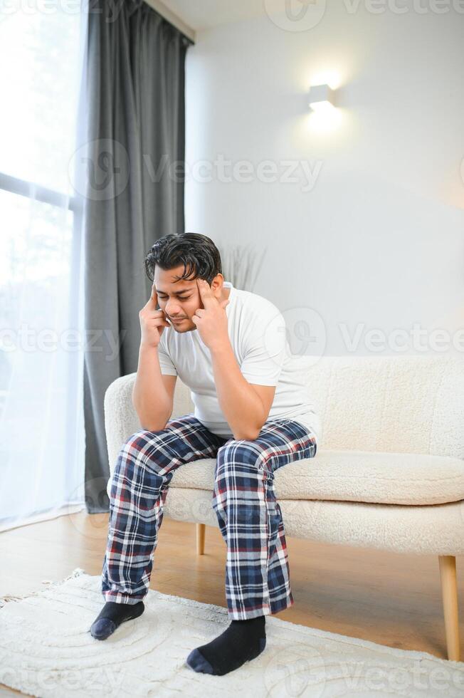 portret van jong Indisch vent lijden van migraine Bij huis, oostelijk Mens gevoel onwel, aanraken zijn tempels met Gesloten ogen, lijden acuut hoofdpijn terwijl zittend Aan bankstel foto