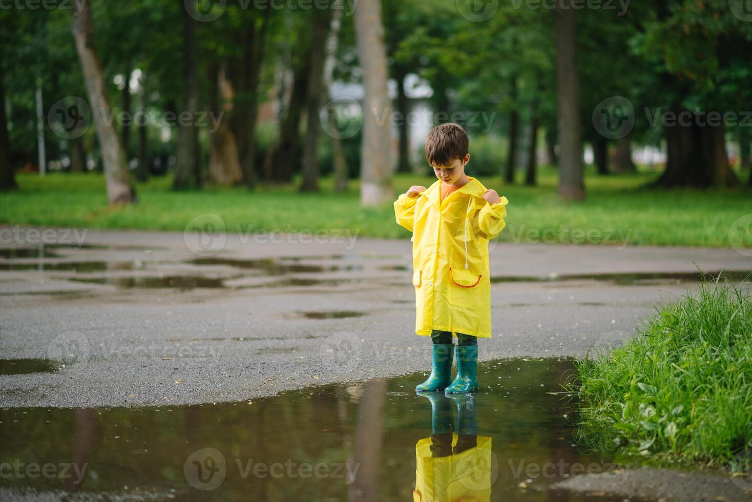 weinig jongen spelen in regenachtig zomer park. kind met paraplu, waterbestendig jas en laarzen jumping in plas en modder in de regen. kind wandelen in zomer regen buitenshuis pret door ieder het weer. gelukkig jeugd. foto