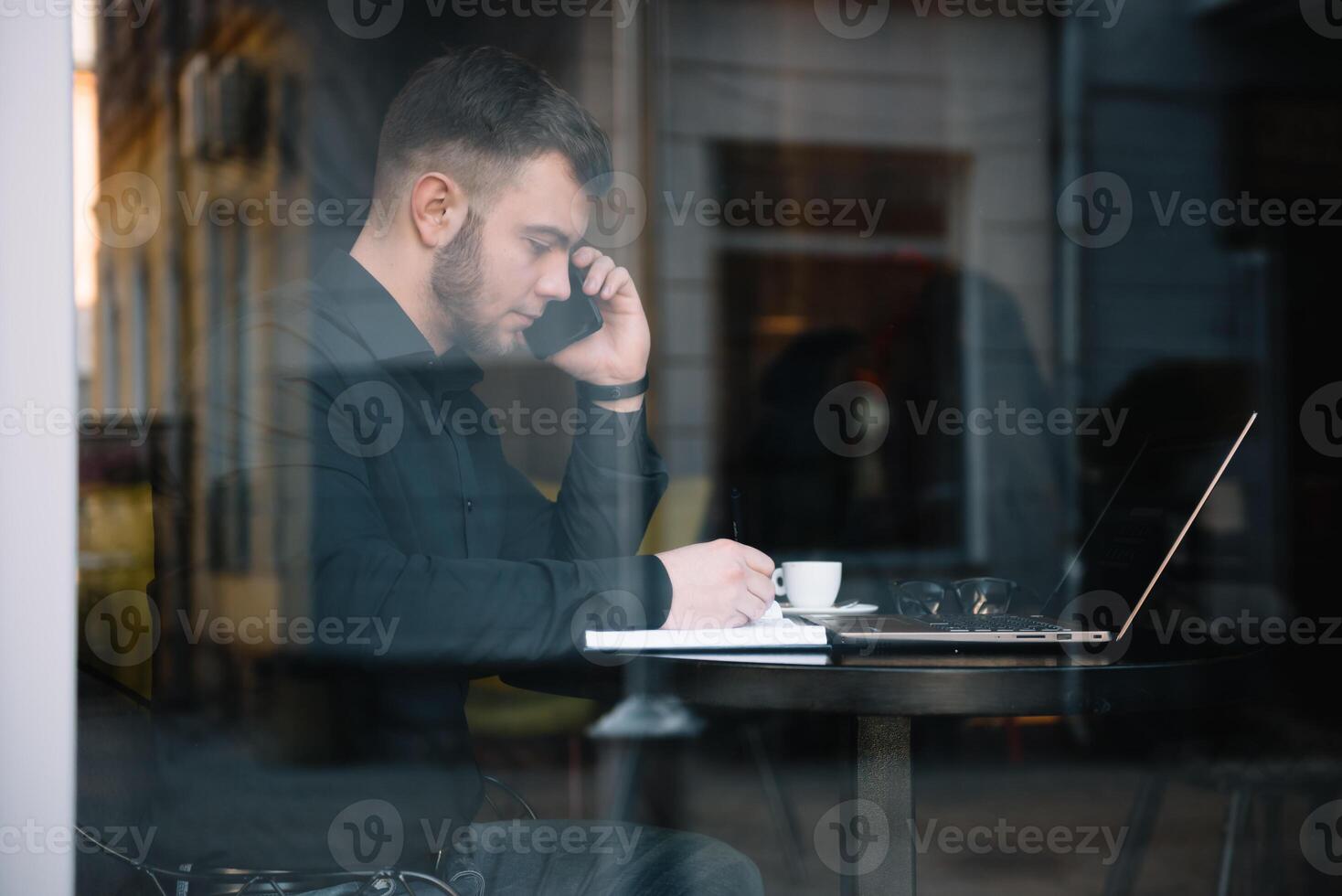 jong zakenman pratend Aan mobiel telefoon terwijl werken Aan laptop in cafe. foto