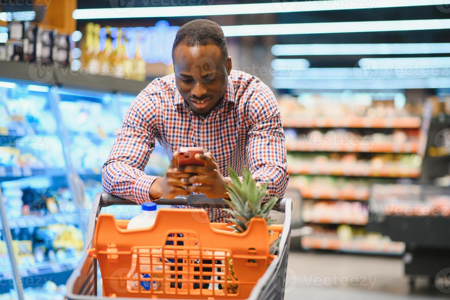 gefocust jong Afrikaanse Amerikaans mannetje klant in gewoontjes kleren berichten Aan mobiel telefoon terwijl staand in de buurt schappen in kruidenier winkel foto