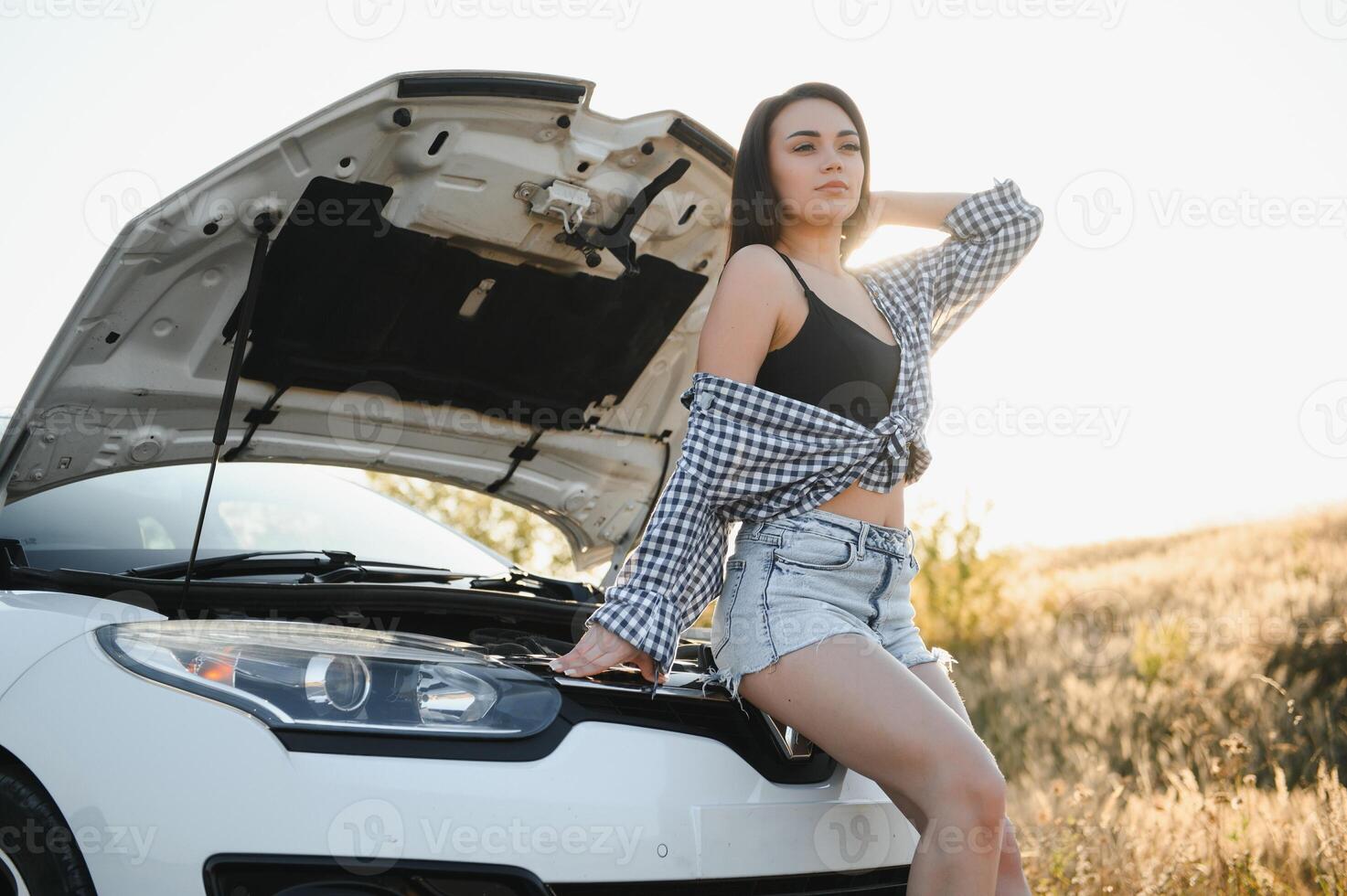 aantrekkelijk slank jong meisje in zomer shorts en overhemd reparaties een gebroken auto. een mooi vrouw staat in de buurt verheven auto kap. foto