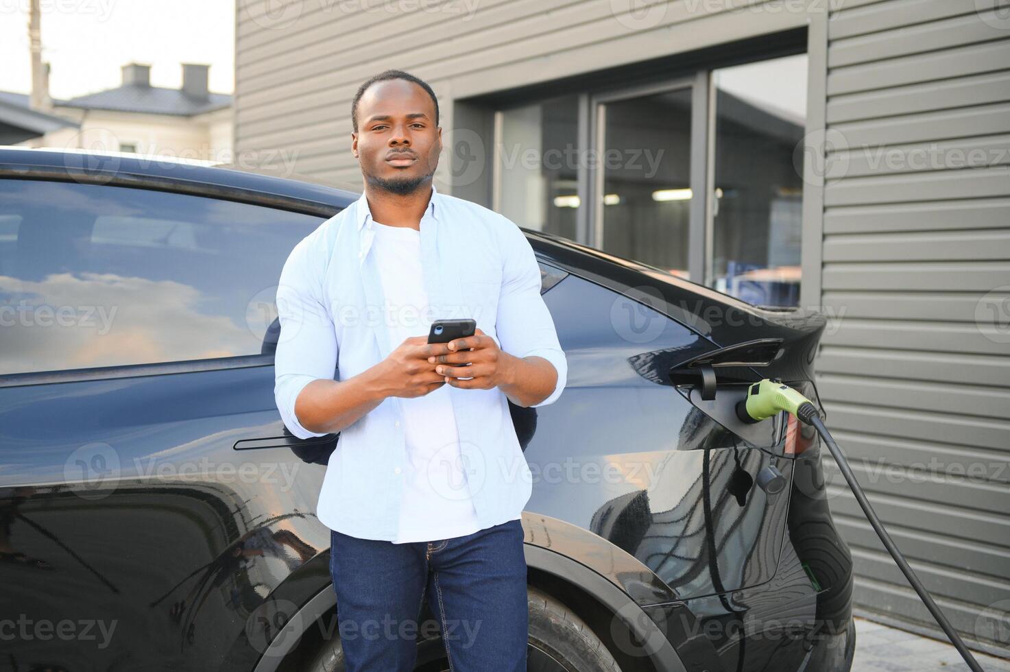 een Afrikaanse Mens is staand in de buurt een elektrisch auto, aan het wachten voor het naar in rekening brengen Bij een opladen station en gebruik makend van de telefoon. foto