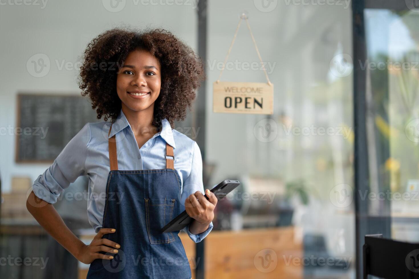 geslaagd Afrikaanse Amerikaans vrouw in schort staand koffie winkel deur. gelukkig klein bedrijf baasje. glimlachen portret van ondernemer staand met kopiëren ruimte. foto