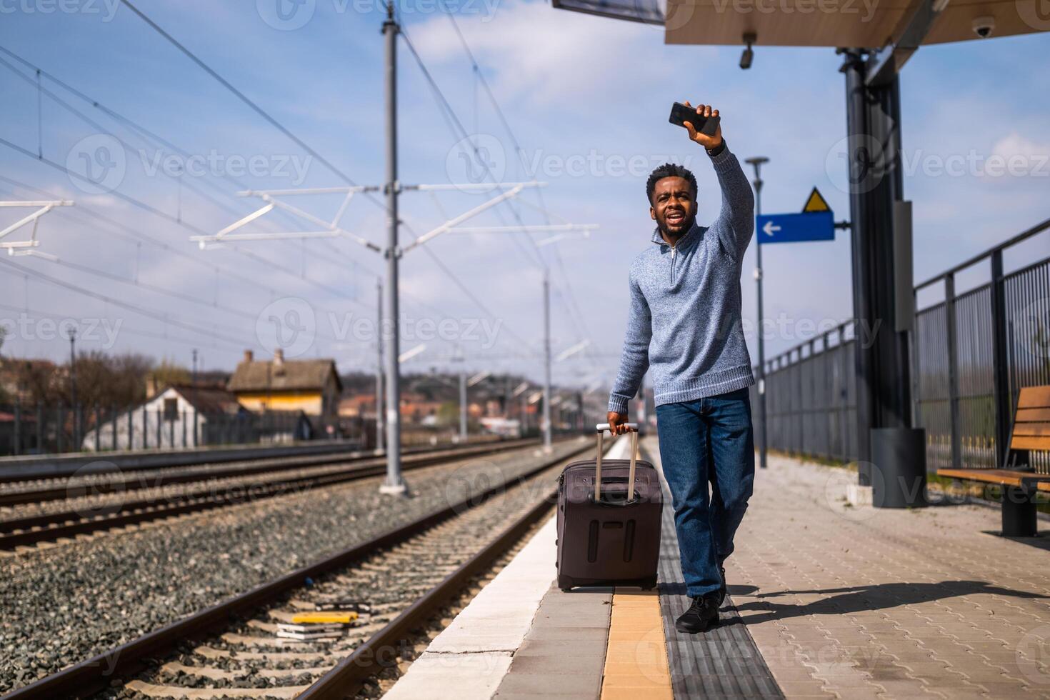 Mens golvend naar een weggaan trein en rennen langs spoorweg station met koffer en mobiel telefoon. foto