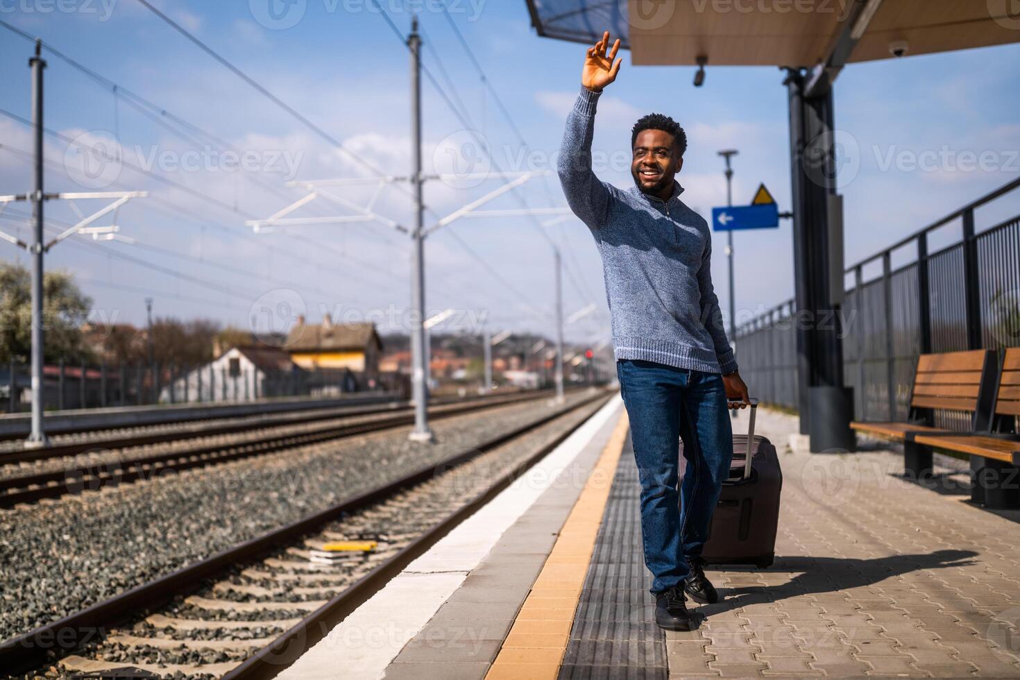 gelukkig Mens met koffer staand en golvend Aan een spoorweg station. foto
