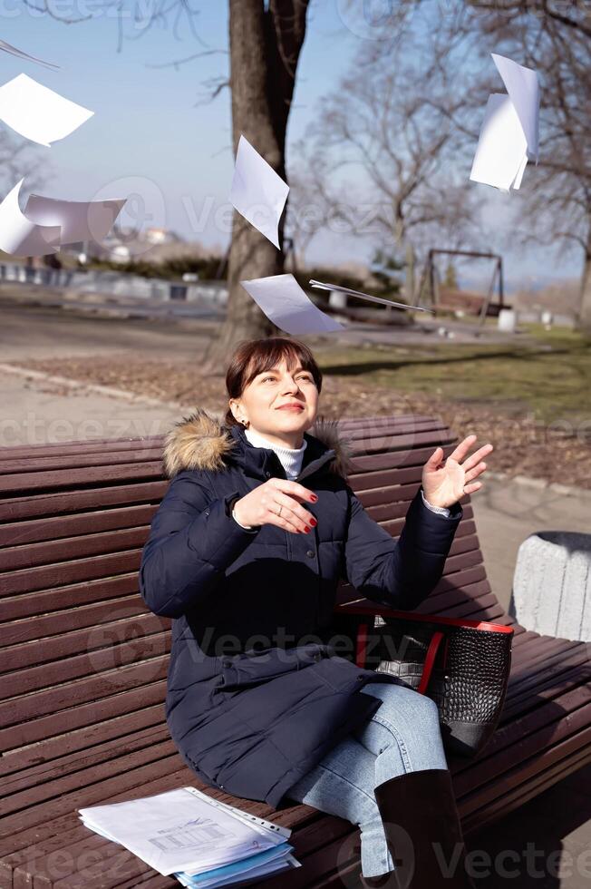 een jong vrouw accountant zit Aan een bank in de park en met een glimlach Aan haar gezicht gooit omhoog rapportage documenten, rapportage periode foto