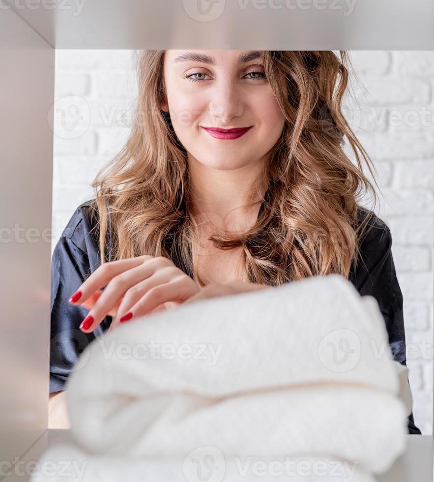 Glimlachende jonge vrouw die een stapel handdoeken schikt op een plank op witte bakstenen achtergrond foto