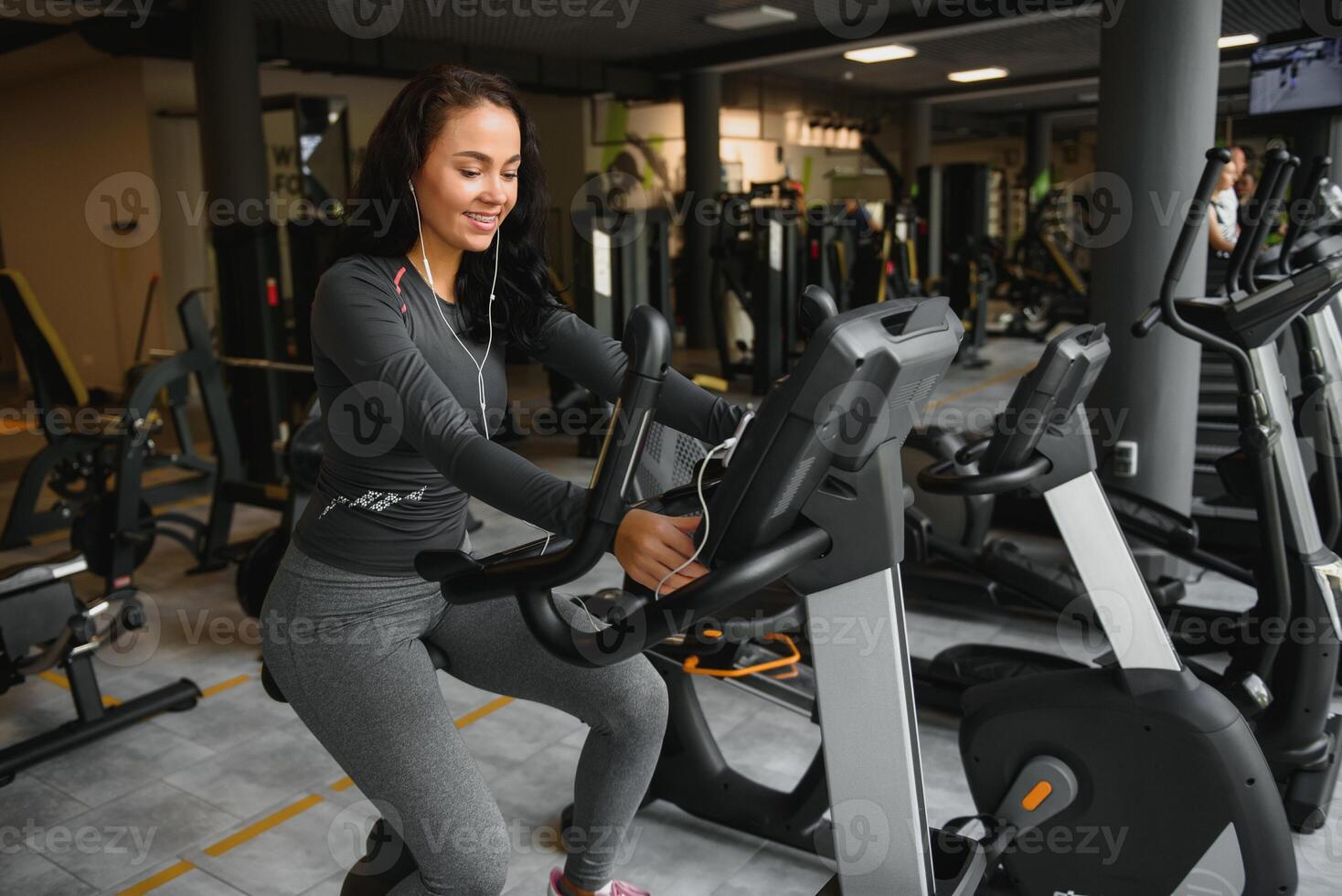 jong vrouw met hoofdtelefoons aan het doen opdrachten Aan schrijfbehoeften fiets in een Sportschool of geschiktheid centrum. jong sportief vrouw in Sportschool luister muziek- van smartphone. Dames aan het doen cardio opdrachten foto