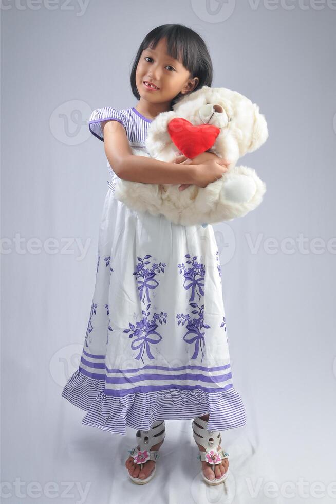 een Indonesisch weinig meisje vervelend modieus jurk en knuffelen een pop met modellering houding. foto