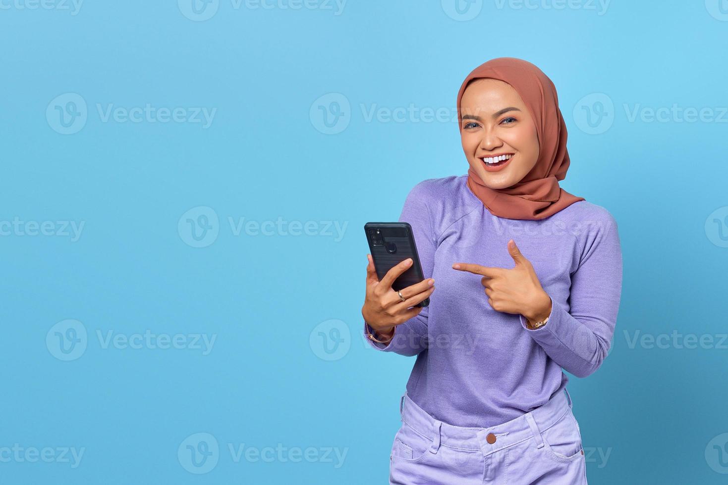 portret van lachende jonge Aziatische vrouw wijzende vinger op een mobiele telefoon op blauwe achtergrond foto