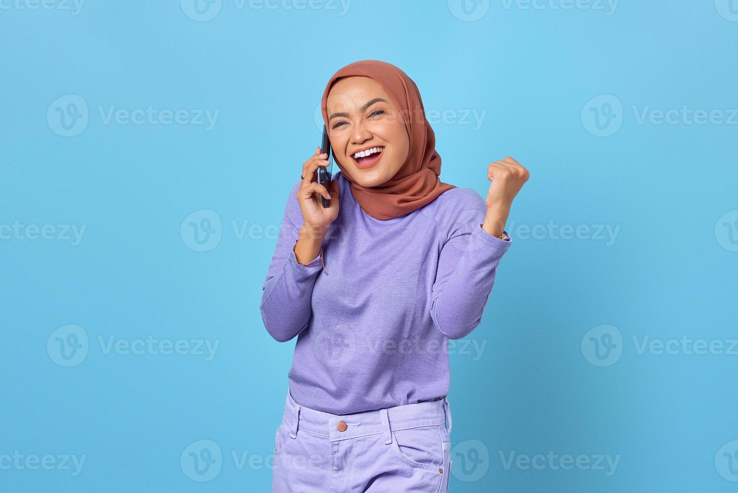 vrolijke jonge aziatische vrouw die op mobiele telefoon praat terwijl ze de overwinning viert op blauwe achtergrond foto
