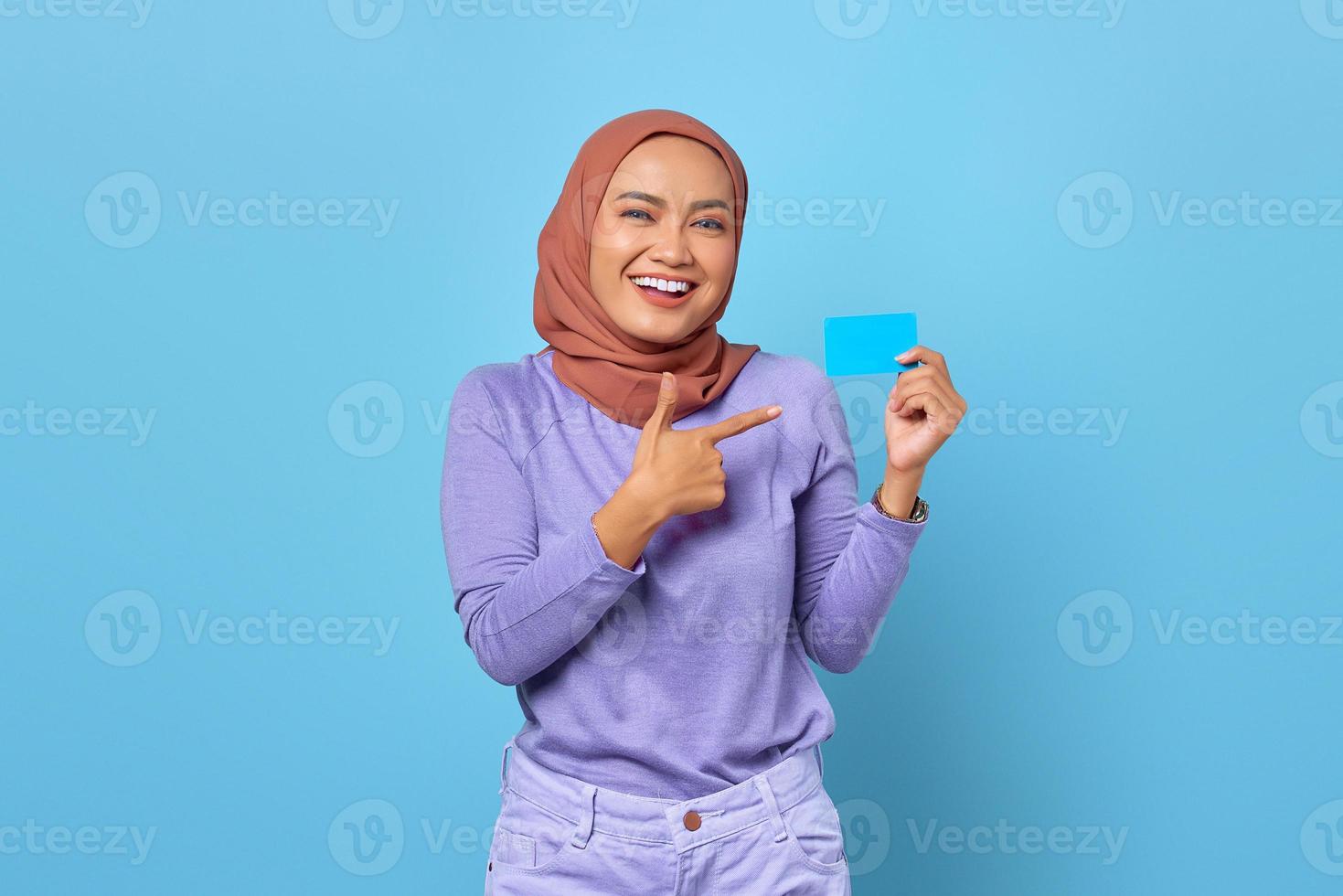 portret van lachende jonge Aziatische vrouw wijzende vinger op een creditcard op blauwe achtergrond foto