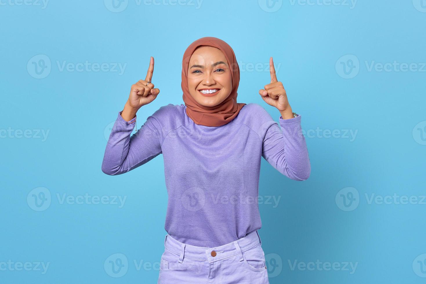 portret van glimlachende jonge aziatische vrouw die met de vinger omhoog wijst op een blauwe achtergrond foto