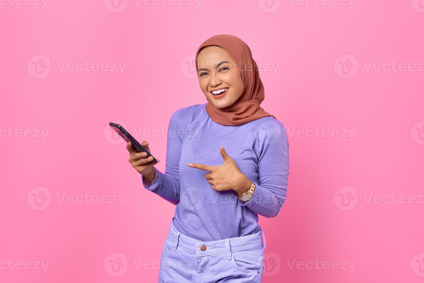 portret van lachende jonge Aziatische vrouw wijzende vinger op een mobiele telefoon op roze achtergrond foto