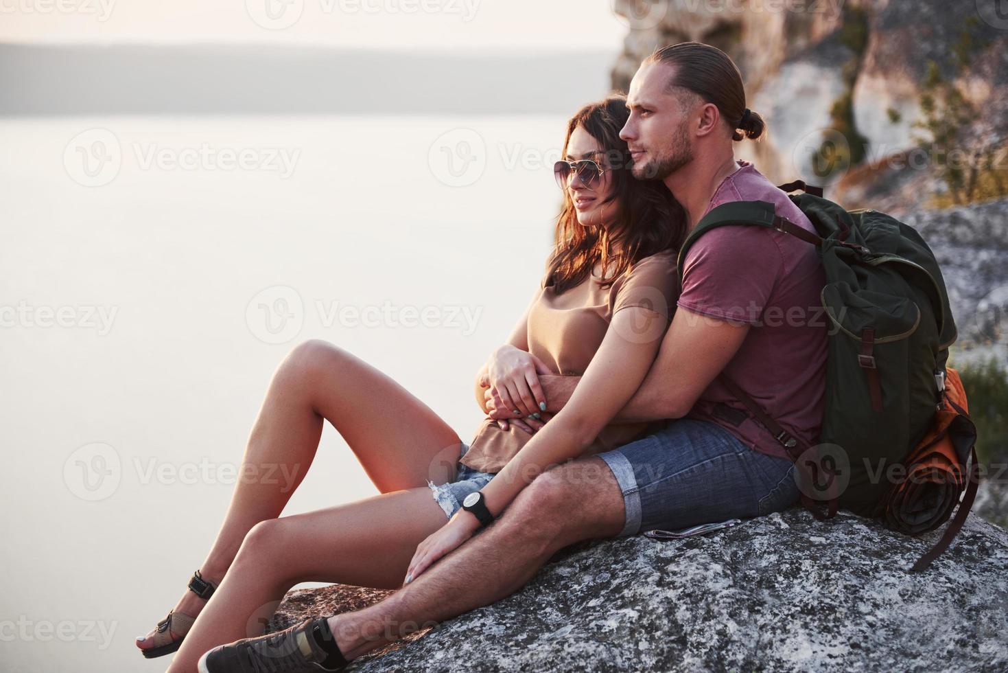 knuffelend paar met rugzak zittend op de top van rock mountain genieten van uitzicht kust een rivier of meer. reizen langs bergen en kust, vrijheid en actief levensstijlconcept foto