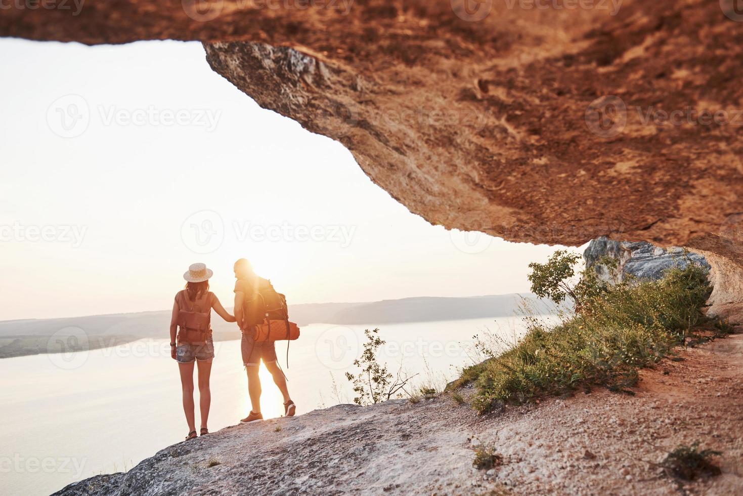 twee toeristische mannen en vrouwen met rugzakken staan naar de top van de rots en genieten van de zonsopgang. reizende bergen en kust, vrijheid en actief levensstijlconcept foto