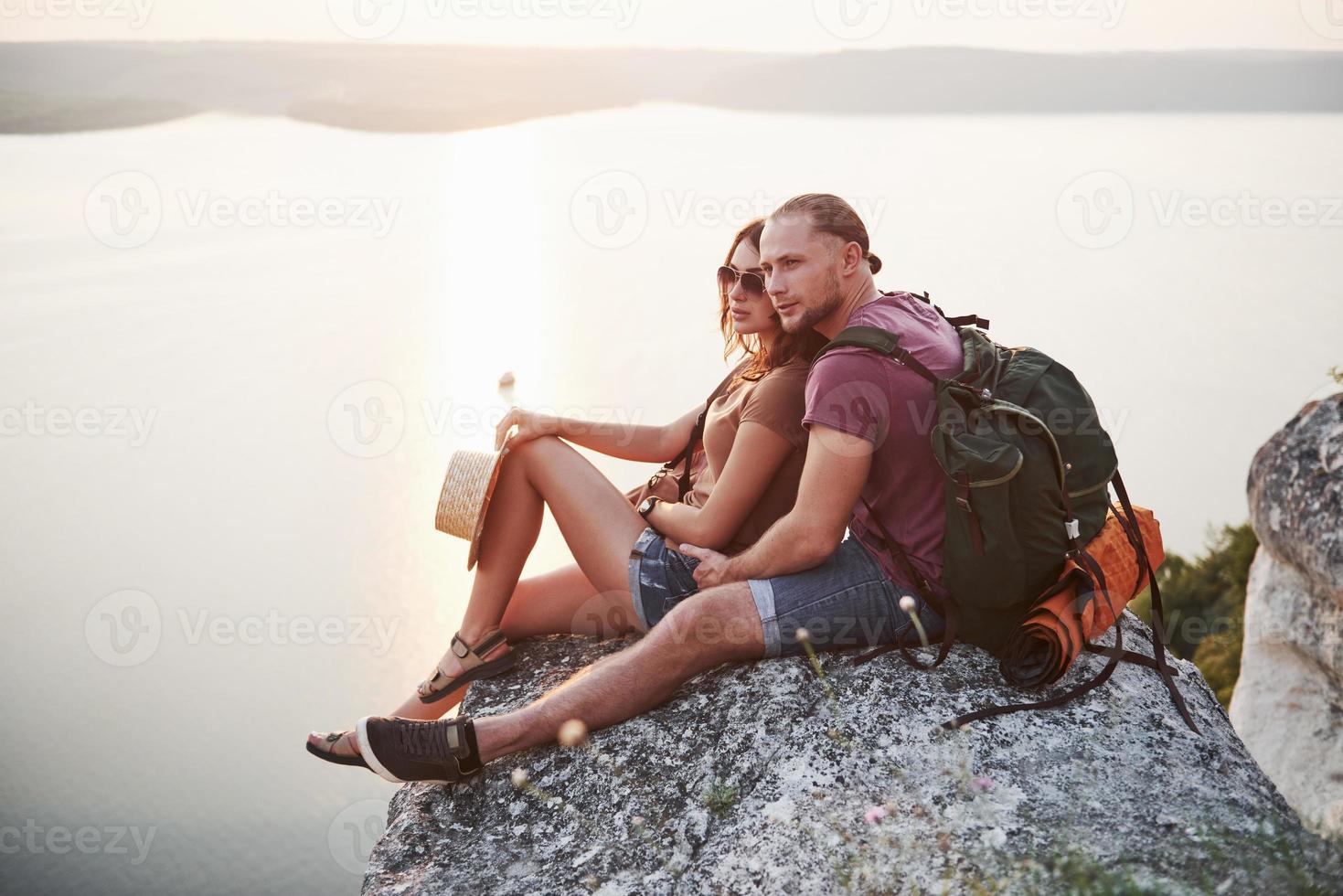 knuffelend paar met rugzak zittend op de top van rock mountain genieten van uitzicht kust een rivier of meer. reizen langs bergen en kust, vrijheid en actief levensstijlconcept foto