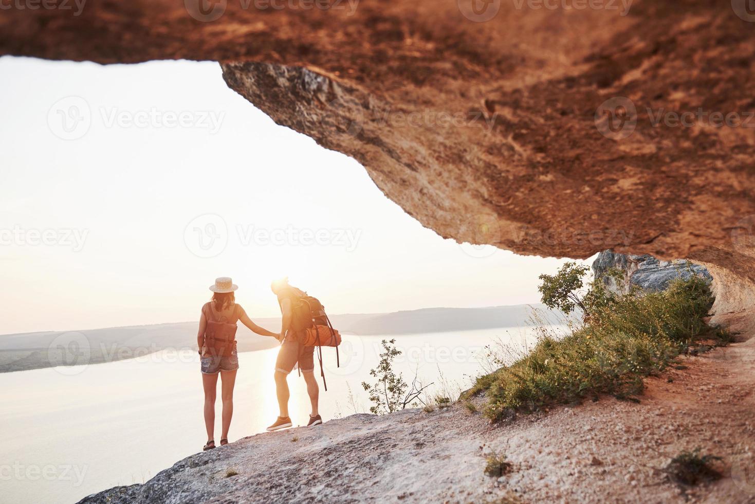twee toeristische mannen en vrouwen met rugzakken staan naar de top van de rots en genieten van de zonsopgang. reizende bergen en kust, vrijheid en actief levensstijlconcept foto