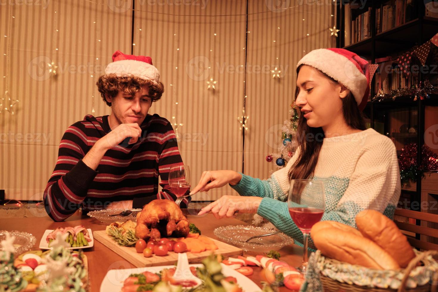 een jong stel geniet van eten aan tafel met speciale gerechten, vriendin die geroosterde kalkoen snijdt in de eetkamer van huis, versierd met ornamenten, kerstfestival en nieuwjaarsfeest. foto