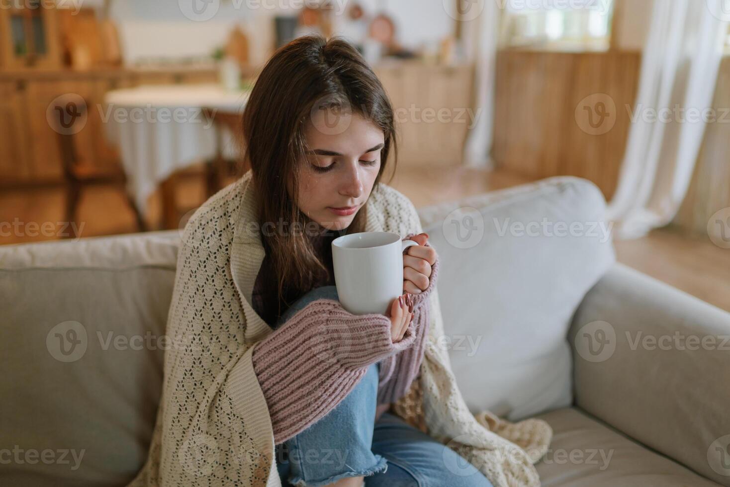 jong vrouw vervelend een gebreid trui zittend Bij huis Aan de sofa verpakt in een plaid drinken heet koffie of thee. bevroren vrouw zullen warm omhoog in een knus leven kamer. foto