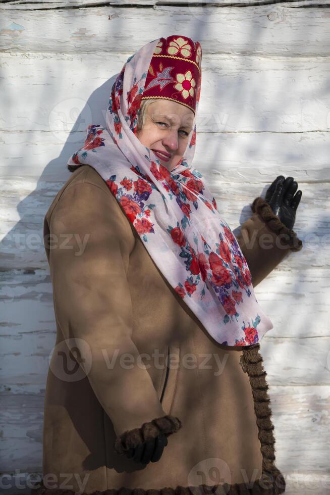 een mooi ouderen Russisch vrouw, dame, in een nationaal hoofdtooi, kokoshnik en schapenvacht jas, tegen een houten achtergrond. foto