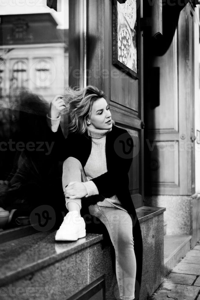 vrouw in zwart jas Aan steen richel. een vrouw met door de wind geveegd haar- zit Aan een steen richel, gekleed in een elegant zwart jas. de beeld vangt een moment van overpeinzing temidden van de elementen foto