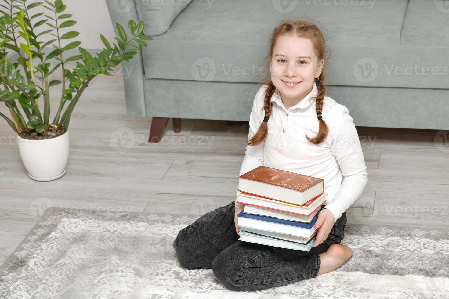 schattig weinig meisje zittend Aan de verdieping Bij huis met een stack van boeken in haar handen foto