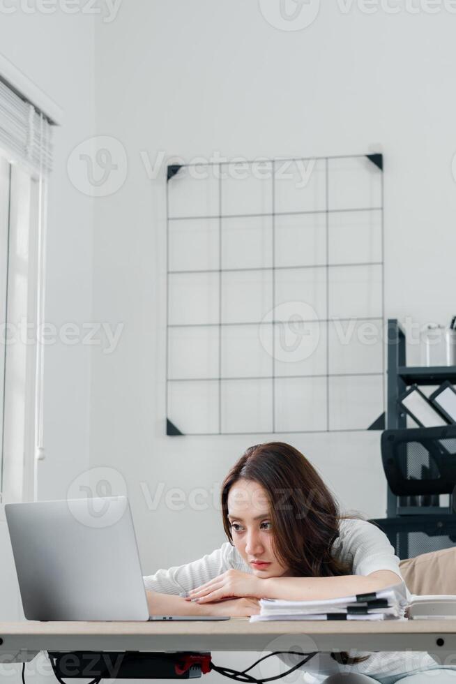 jong vrouw verschijnt overweldigd en gestrest, resting haar hoofd Aan haar armen Bij haar werk bureau met een laptop en papieren. foto