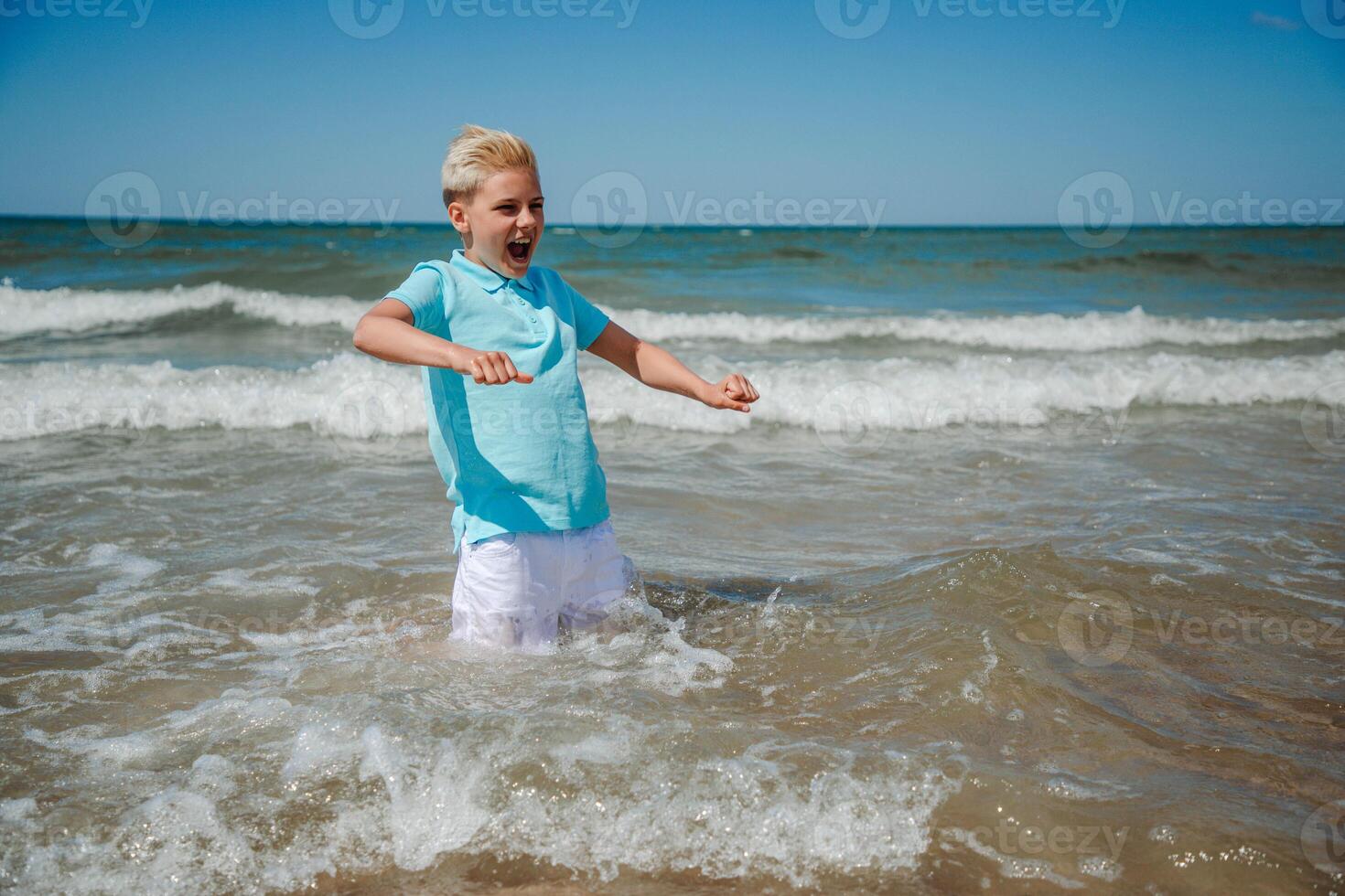 knap gelukkig tiener jongen van Europese uiterlijk met blond haar- in wit korte broek, en een blauw t-shirt zittend en spatten in de zee, en looks naar de camera. zomer familie vakantie concept.zomer reizen concept.kopie ruimte. foto