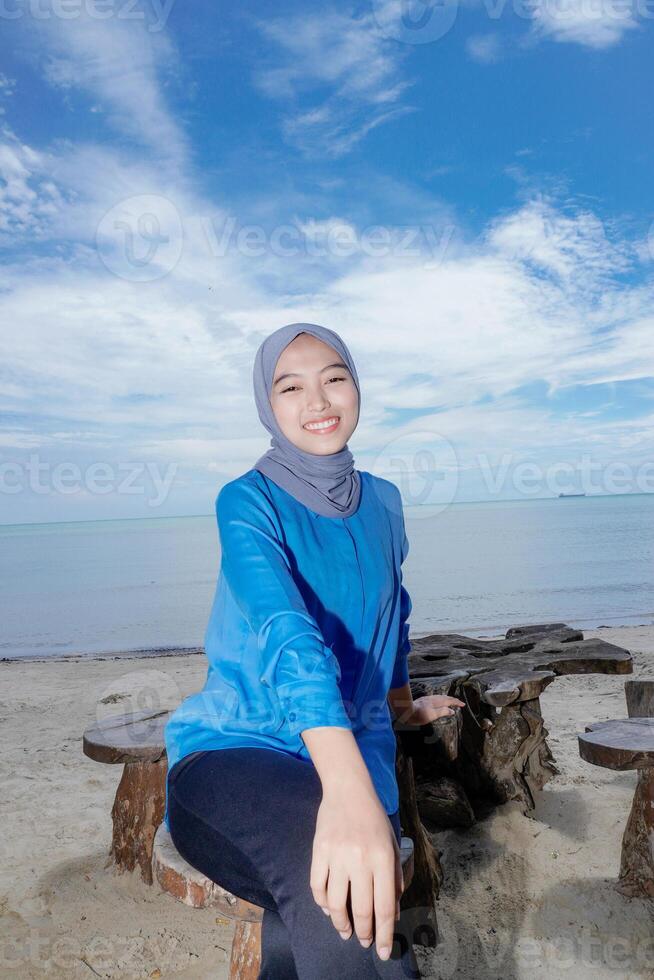 mooi moslim vrouw is zittend Aan de strand op zoek Bij de camera, de achtergrond is een Doorzichtig lucht met leeg fotokopie ruimte foto