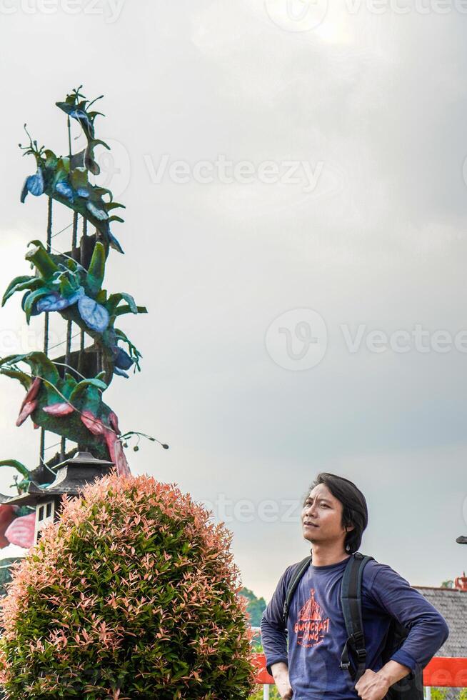 Javaans Mens is staand met een rugzak Aan zijn schouders tegen een bewolkt lucht met vrij ruimte voor fotokopiëren. foto