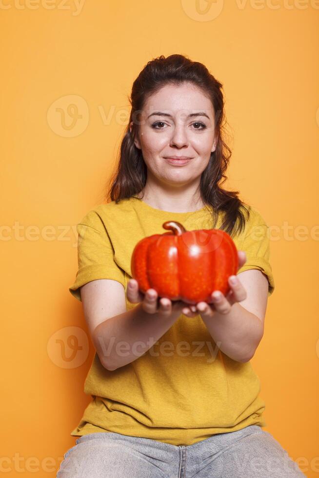 portret van Kaukasisch vrouw aangrijpend een klok peper en op zoek in camera staand in studio met oranje achtergrond. jong dame hebben gezond eetpatroon, aan het eten groenten. biologisch vegetarisch concept. foto