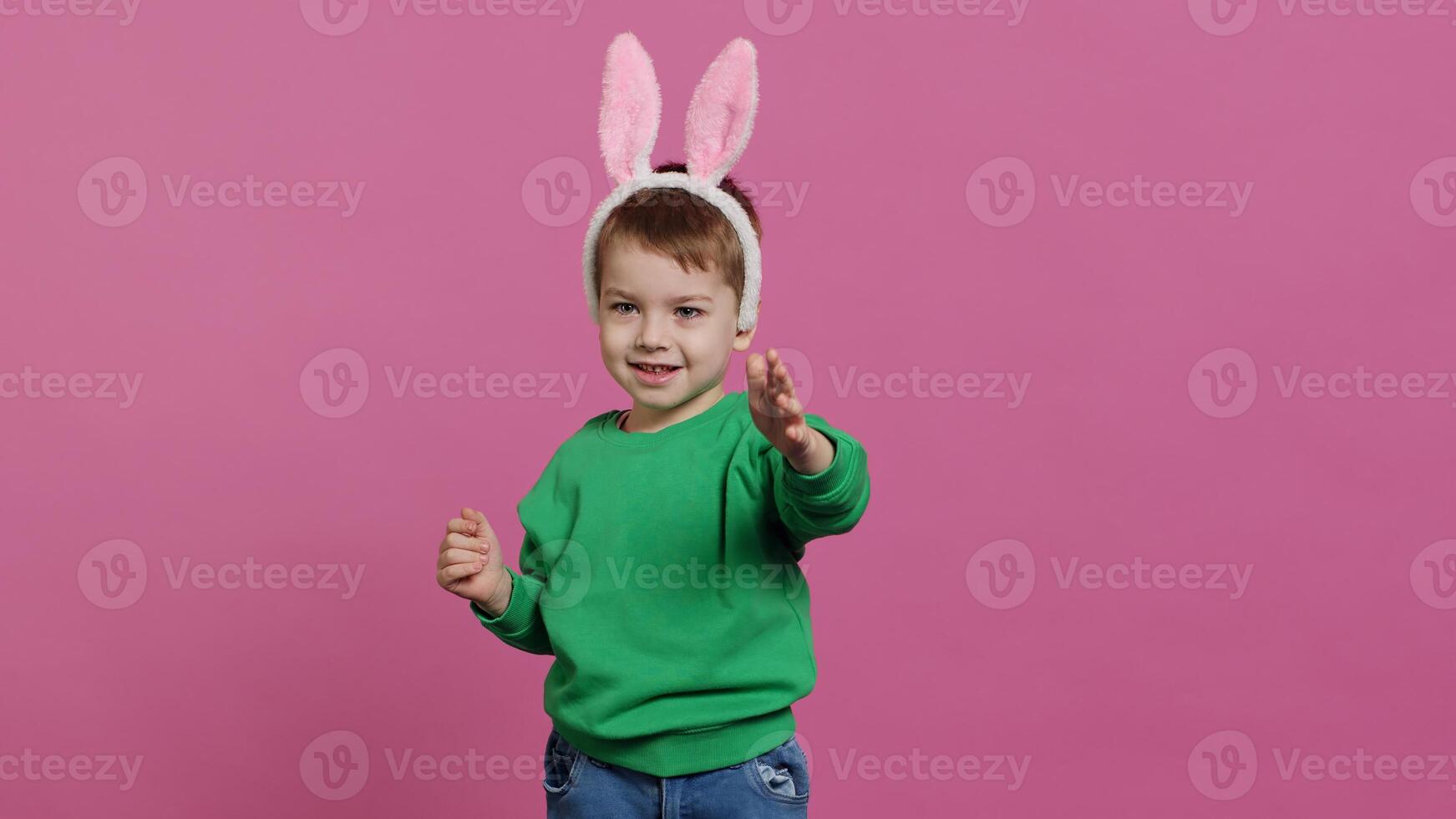 schattig weinig jongen vervelend pluizig konijn oren in studio en zwaaien, wezen aanbiddelijk tegen roze achtergrond. glimlachen jong kind wezen opgewonden over Pasen vakantie feest, kinderjaren onschuld. camera a. foto