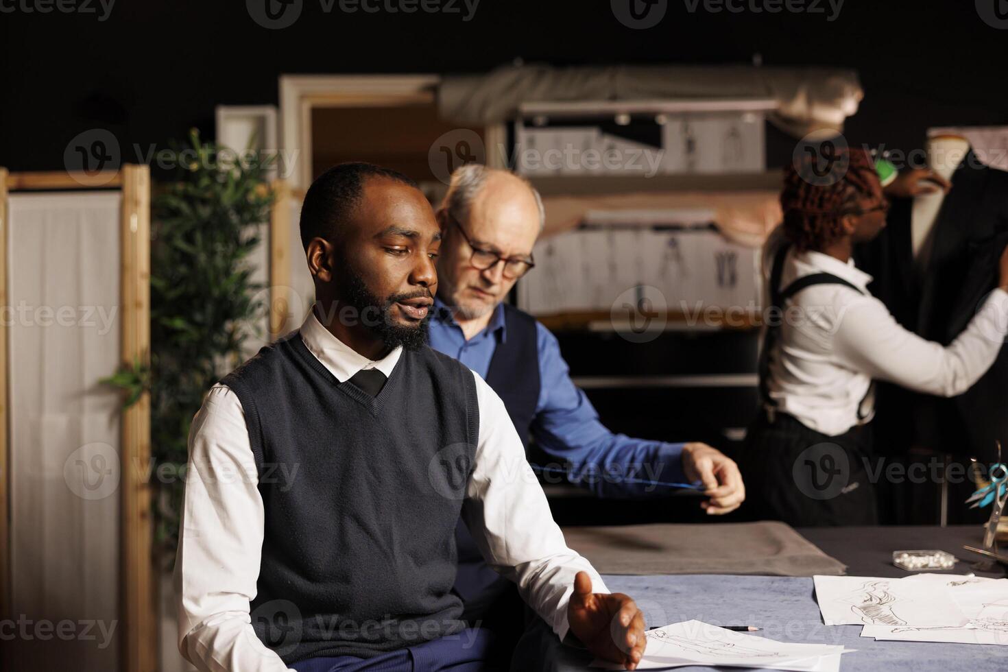 meester kleermaker nemen cliënt metingen terwijl nauwkeurig naaister leerling doet handwerk Aan pak in achtergrond. heer krijgen Op maat gemaakt kleding gedaan door professioneel couturiers team foto