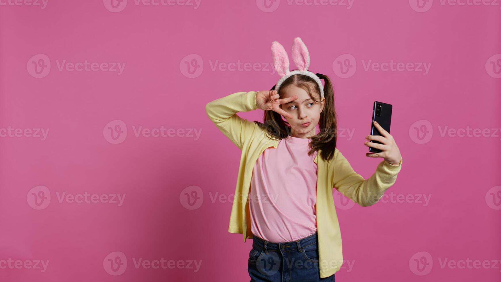 zelfverzekerd gelukkig jong meisje nemen foto's Aan smartphone webcam, gevoel schattig met haar vlechten en konijn oren. blij glimlachen kleuter duurt afbeeldingen en voor de gek houden in de omgeving van in studio. camera b. foto