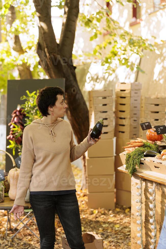 jong vrouw klant met een aubergine in hand, inkoop plaatselijk gegroeid kleurrijk vers natuurlijk produceren. vrouw klant staand Bij boeren markt kraam, klaar naar kopen vers geoogst vegetarisch. foto