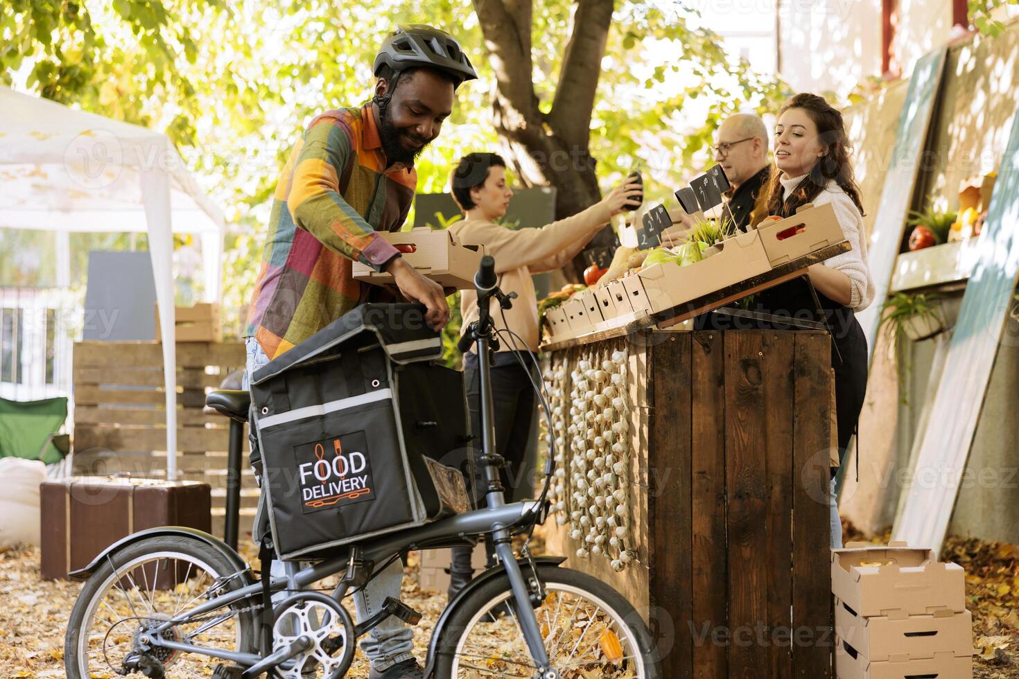 zwart mannetje koerier zetten doos van biologisch producten in rugzak voor leveren bestellen naar klanten, gebruik makend van fiets. jong vrolijk postbode werken Aan boeren markt groenten Verzenden. foto