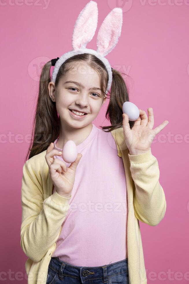 jong schattig meisje poseren met vertrouwen in voorkant van camera, presenteren haar geschilderd roze eieren voor Pasen feest. weinig schoolmeisje vervelend konijn oren glimlachen in studio, shows handgemaakt decoraties. foto