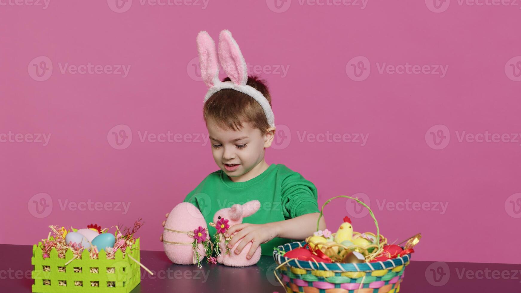 blij jong kind spelen in de omgeving van met feestelijk geschilderd decoraties, tonen een konijn speelgoed- en een roze ei in voorkant van camera. glimlachen klein jongen met konijn oren hebben pret met ornamenten. camera a. foto