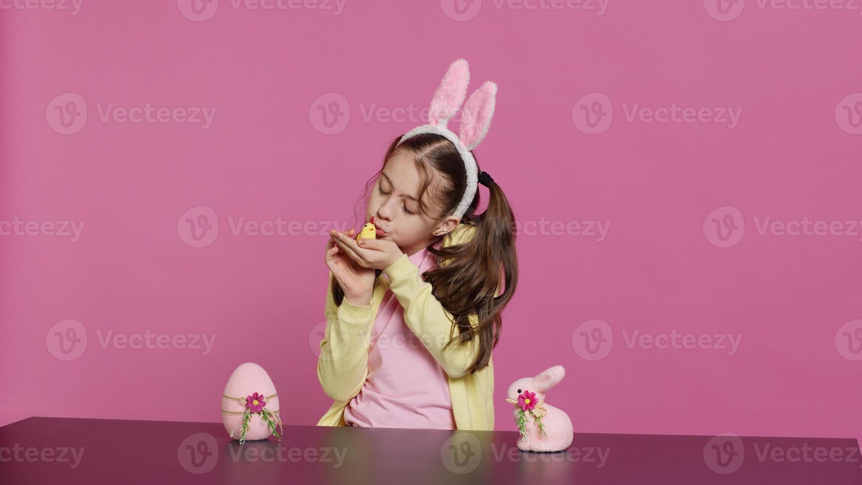 blij weinig meisje spelen met feestelijk Pasen decoraties in studio, creëren regelingen met een kuiken, konijn en ei. glimlachen schattig kleuter met konijn oren tonen kleurrijk ornamenten. camera a. foto