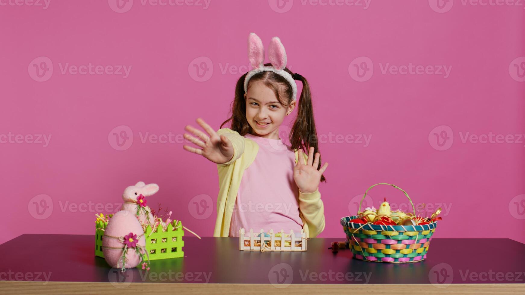 blij schoolmeisje met konijn oren golvend Hallo in voorkant van camera, zittend Bij een tafel met feestelijk decoraties en regelingen voor Pasen zondag viering. jong opgewonden kind. camera b. foto