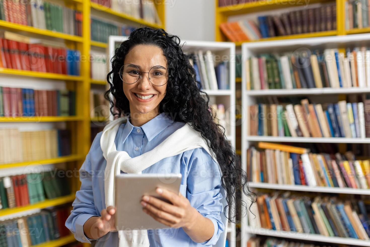 een vrolijk leerling gebruik makend van een digitaal tablet staat in een bibliotheek met een breed glimlach, omringd door schappen vol van boeken. foto