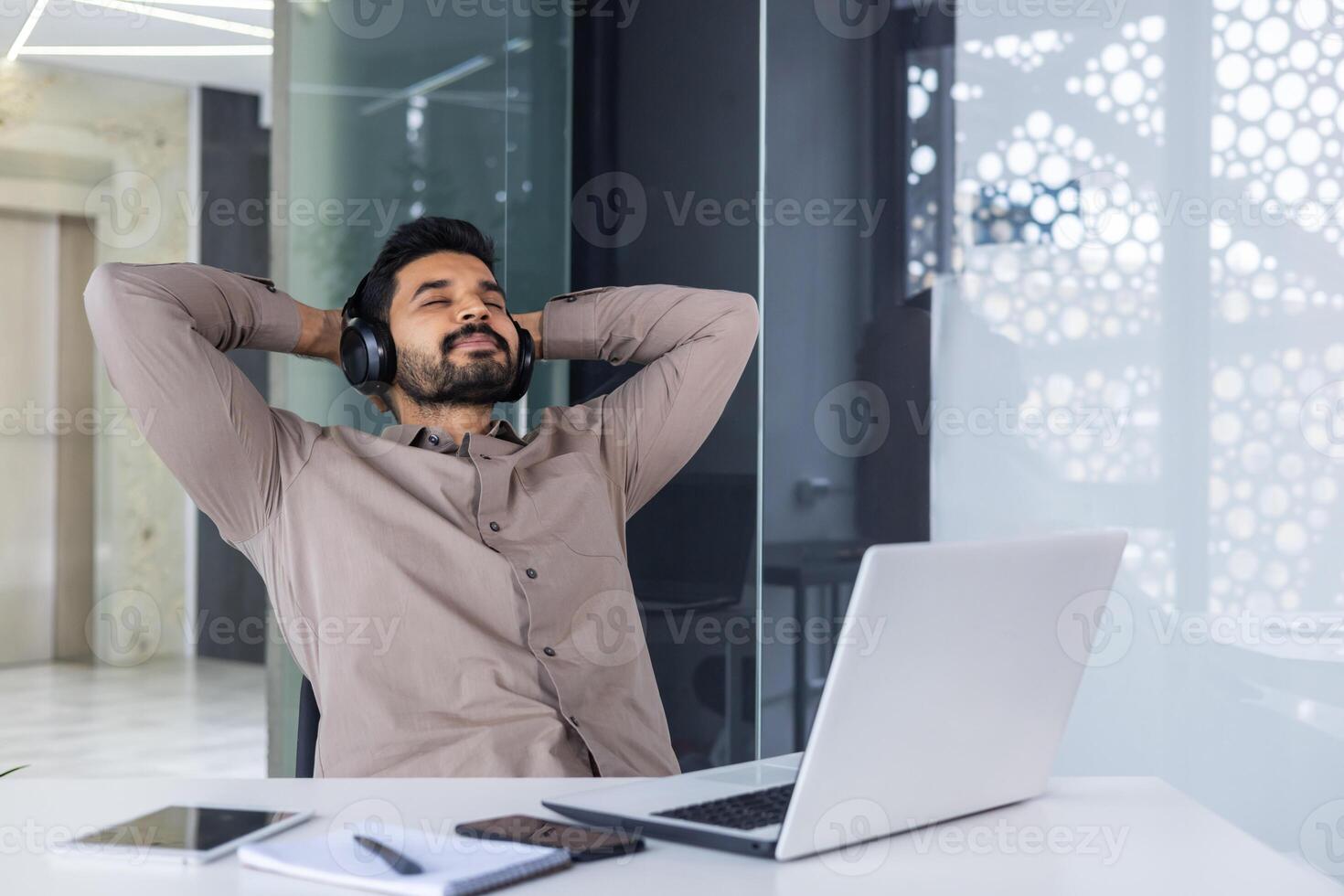 zakenman resting binnen kantoor met zijn handen achter zijn hoofd met ogen Gesloten dommelen, Mens goed gedaan met succes voltooid een project met hoofdtelefoons luisteren naar muziek- en audio boeken podcasten. foto