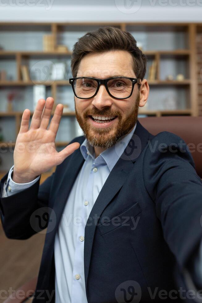 volwassen zakenman in bril en pak glimlachen en golvend Bij de camera, geven een vriendelijk groet van zijn knus kantoor. foto
