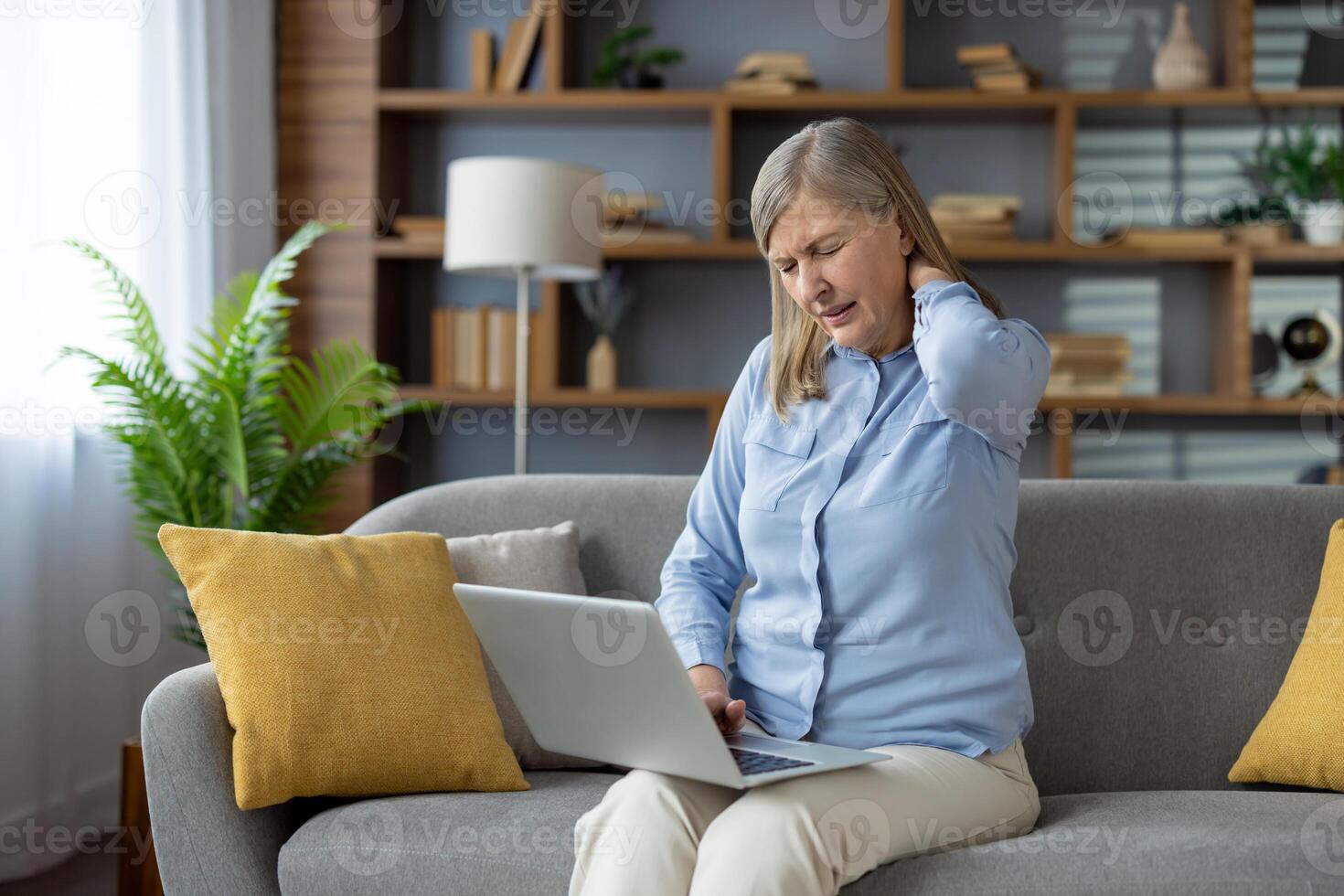 moe volwassen dame in blauw overhemd wrijven gespannen nek terwijl zittend Aan bankstel met laptop Aan poten. overbelast vrouw aanraken terug van hoofd met Gesloten wauw voor verlichtend spanning in spinal spieren. foto