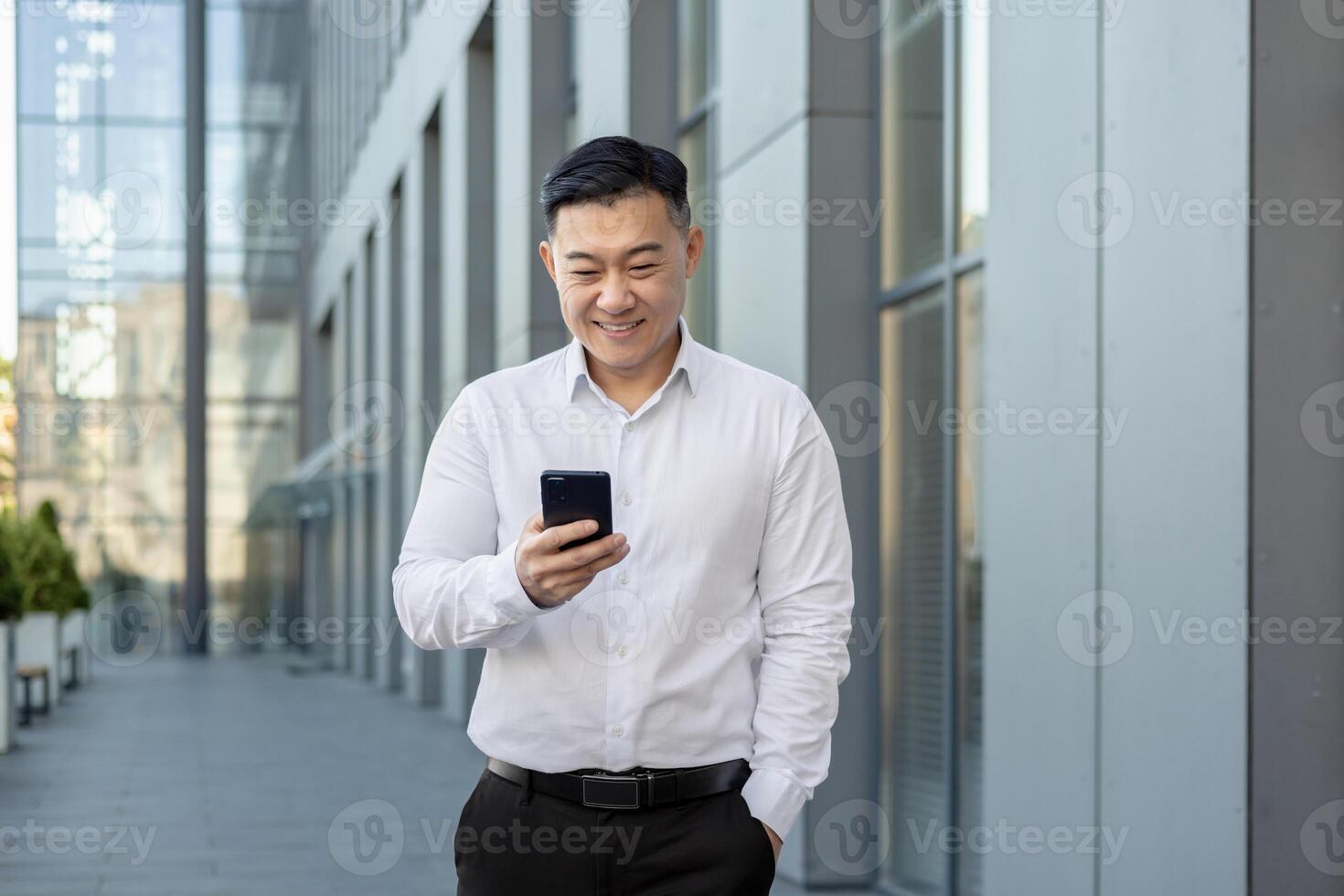 jong professioneel Mens in een wit overhemd verloofd met een telefoon buitenshuis tegen een backdrop van modern architectuur. foto