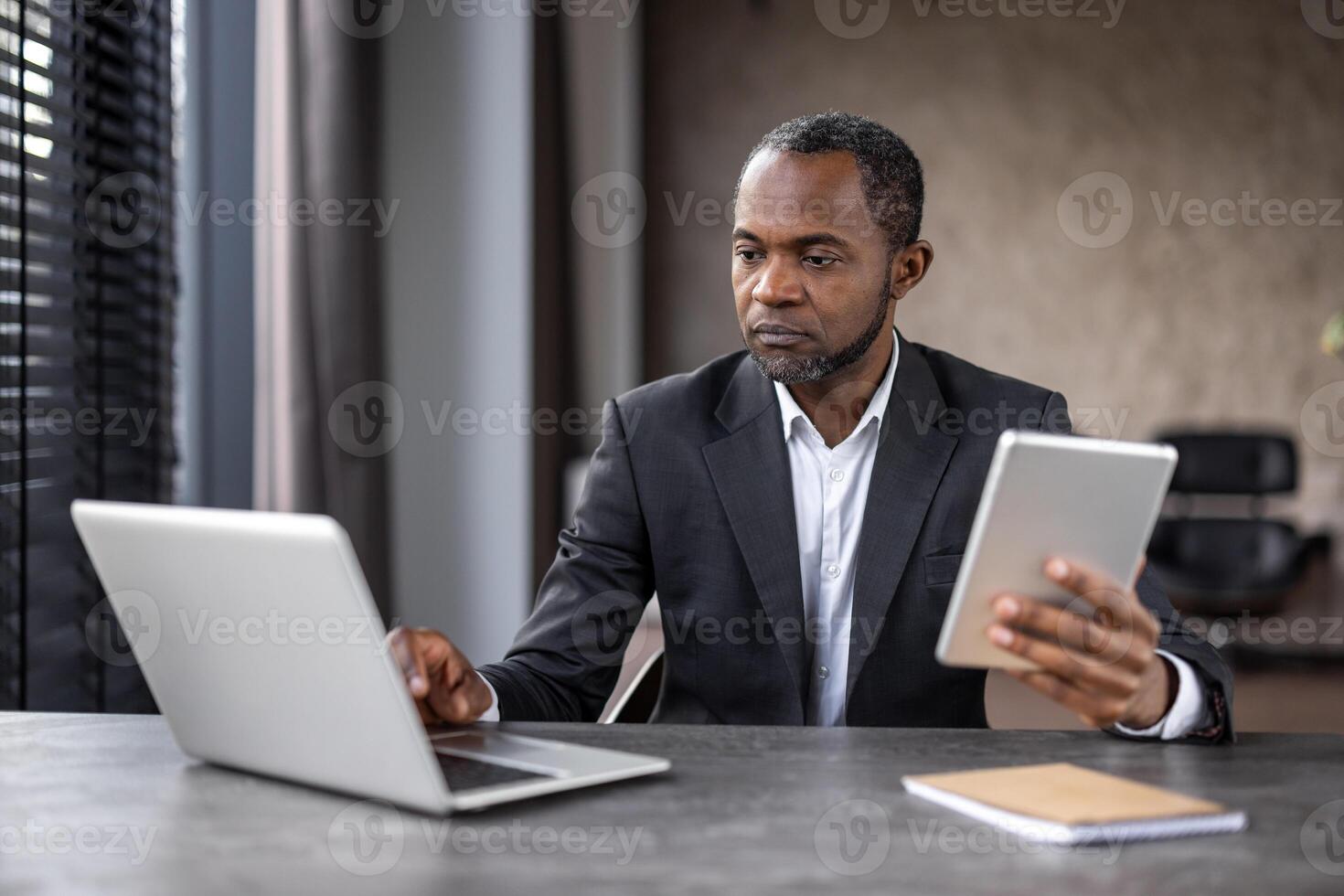 gefocust Afrikaanse Amerikaans zakenman toepassingen een laptop en tablet Bij zijn modern werkruimte, afbeelden toewijding en technologie gebruik in een professioneel instelling. foto