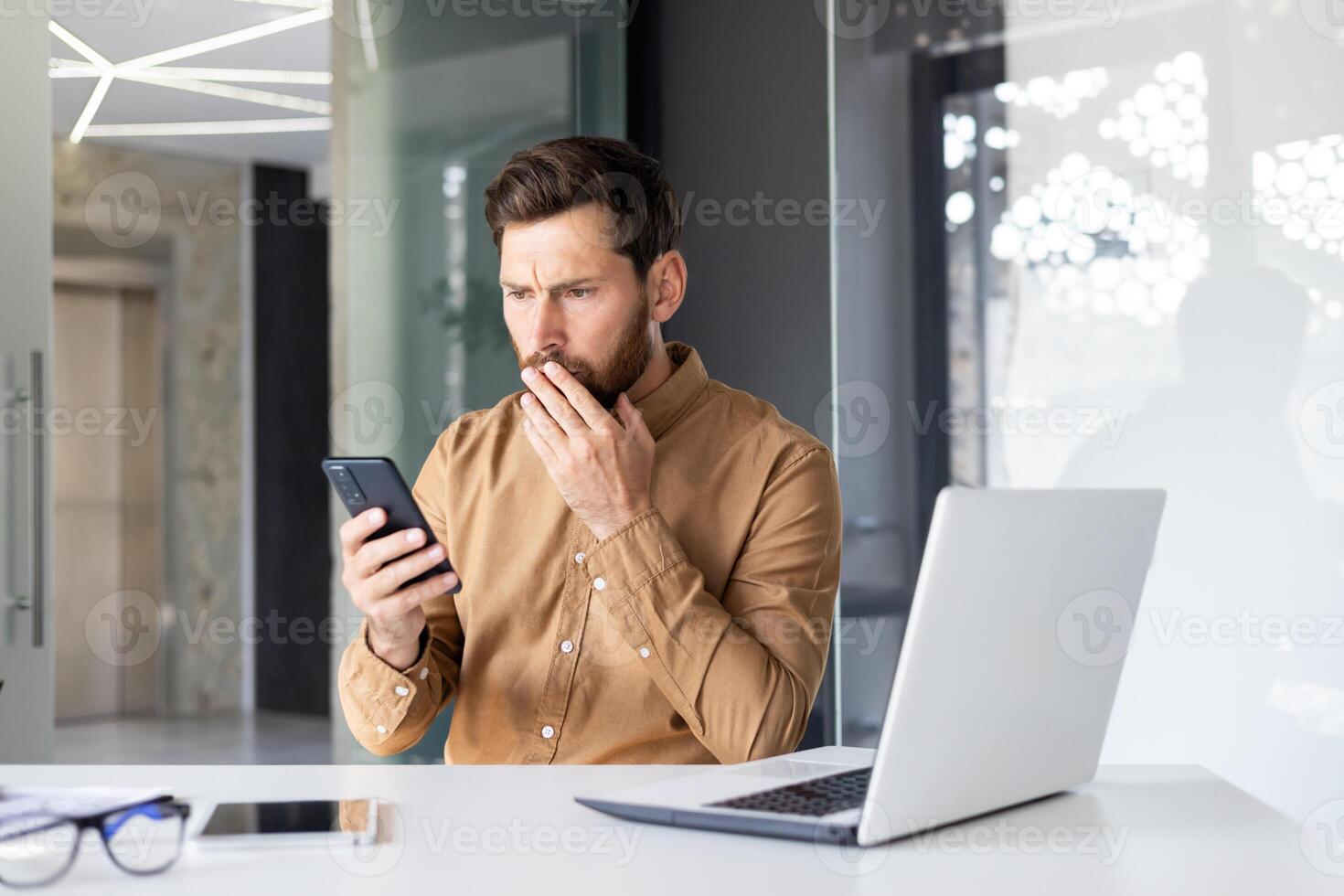 Mens lezing verdrietig slecht nieuws gebruik makend van telefoon, zakenman Bij werkplaats Holding smartphone, geschokt en teleurgesteld binnen kantoor met laptop. foto