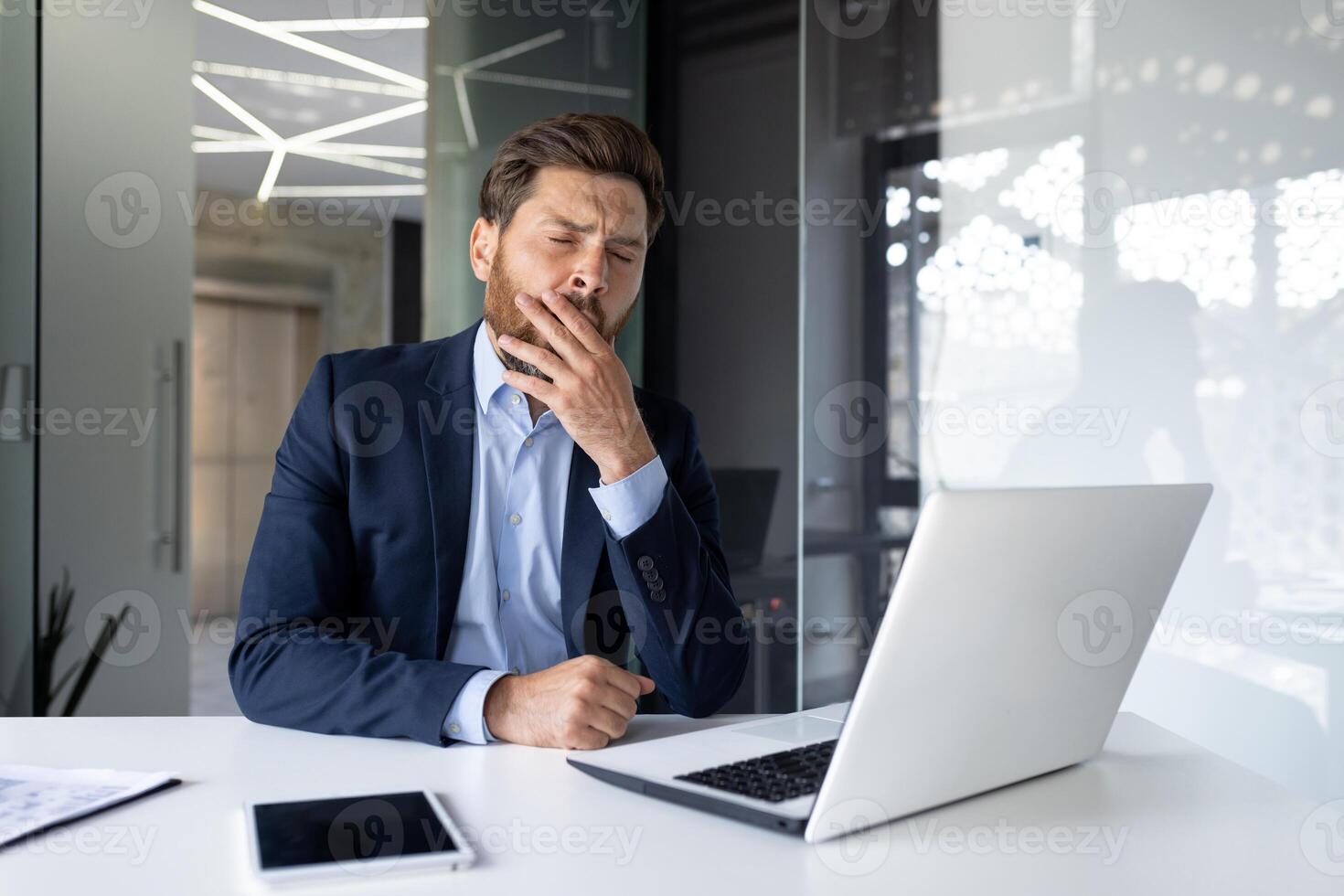 moe jong zakenman Mens in een pak zit in de kantoor Bij de bureau en gaapt, covers zijn mond met zijn hand, voelt slaperig. foto