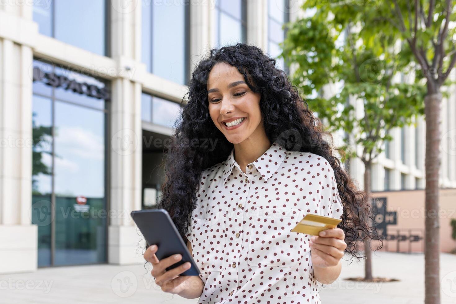 jong mooi vrouw wandelen in de stad, tevreden Latijns Amerikaans vrouw met gekruld haar- houdt telefoon en bank credit kaart in handen, maakt online aankopen gelukkig boeken Diensten en kiest online. foto